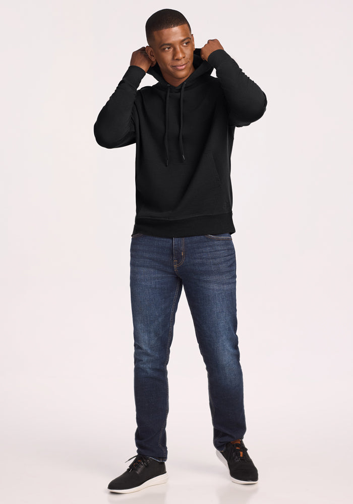 A man stands against a plain background, wearing the Woolx Chase Hoodie in black. He is smiling slightly and adjusting the hood with both hands while paired with blue jeans and black sneakers.