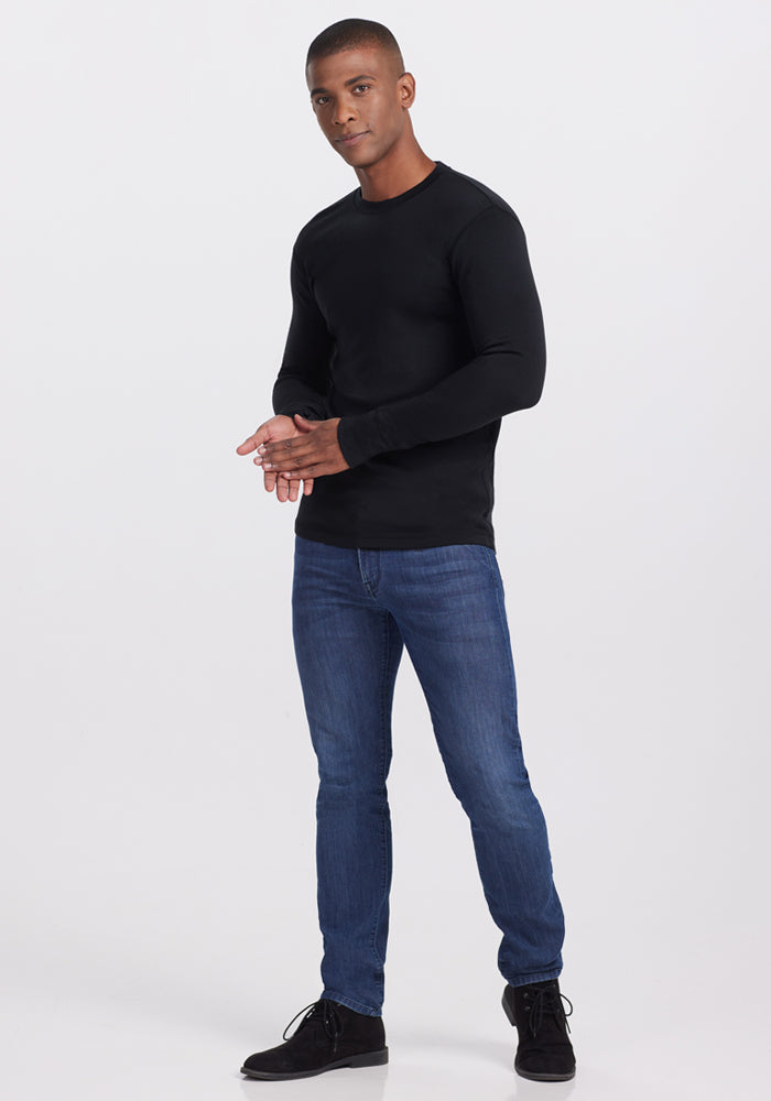 A man stands against a white background wearing the Woolx Glacier Long Sleeve Crew in black, paired with blue jeans and black shoes. He looks forward, hands clasped, with one foot slightly ahead of the other.