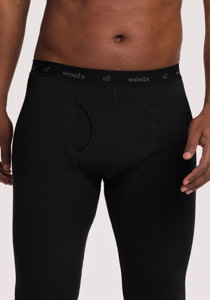 A person is shown from waist to mid-thigh wearing Woolx Arctic Leggings in black, an ultra-heavyweight thermal Merino wool base layer. The black waistband has "woolx" and a small logo repeatedly printed in light gray.