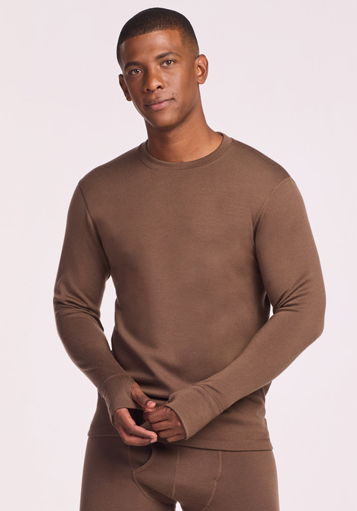 A man stands facing forward with his hands together, wearing the Woolx Glacier Long Sleeve Crew in Bourbon Oak, paired with matching bottoms, against a plain light background. 