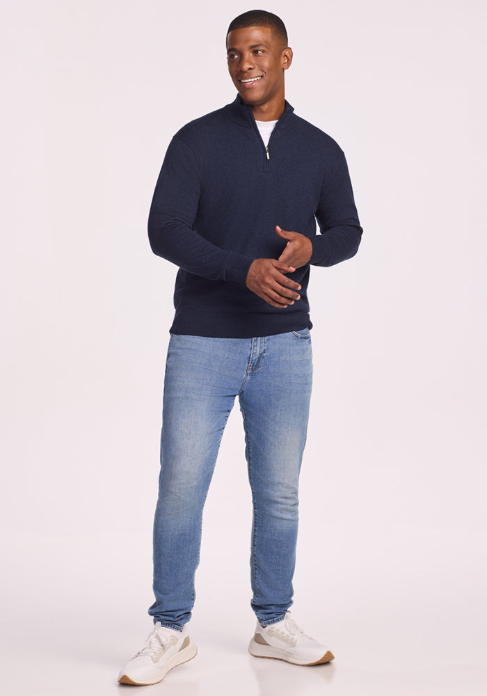 A man smiles wearing the Woolx Enzo Quarter Zip - Storm Cloud as a comfy midweight layer over a white tee, paired with light blue jeans and white sneakers against a plain light background.