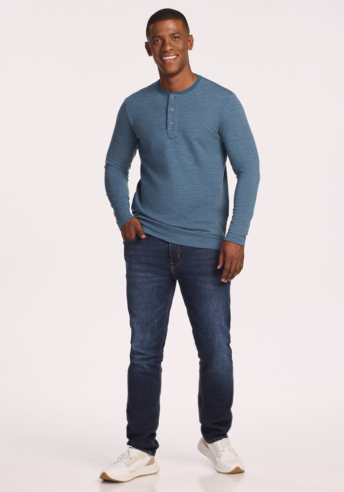 A man smiles, facing forward with one hand in his pocket. He wears the Woolx Jackson Henley in Steel Blue, paired with dark jeans and white sneakers, standing against a plain light background.
