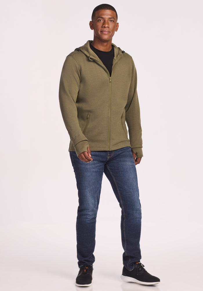 A young man stands facing forward, wearing the Woolx Grizzly Sweatshirt in Winter Moss, paired with dark blue jeans and black sneakers. He has short hair and a neutral expression against a plain white background.