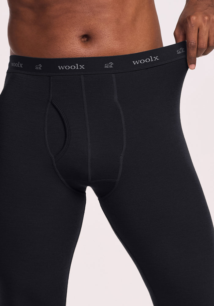 A person wearing Woolx Backcountry Leggings in black, shown from mid-chest to mid-thigh, gently pulls the waistband outward to demonstrate fit and comfort—perfect for cold weather activities.