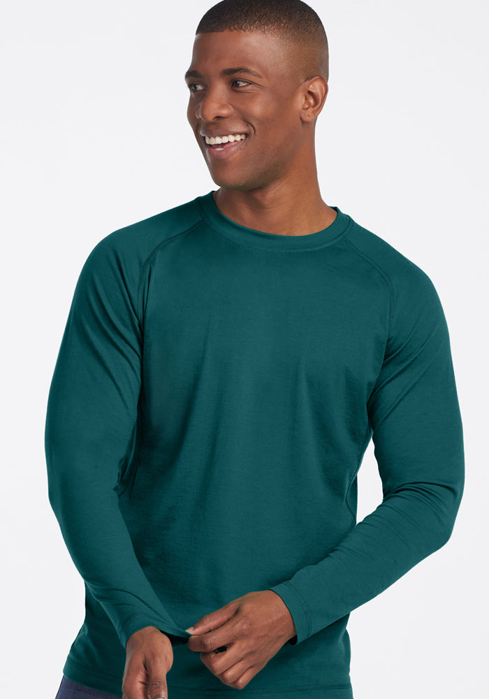 A smiling man models the Woolx Essential Tee in Riverbank Green, a lightweight merino wool long-sleeve shirt, standing against a plain light background and looking slightly to the side with one hand raised mid-gesture.
