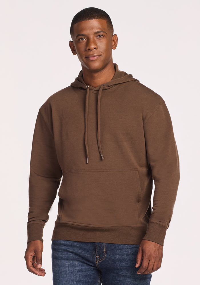 A man stands facing forward against a plain light background, wearing blue jeans and the Woolx Chase Hoodie in Bourbon Oak—a men’s wool hoodie featuring a front pocket and drawstrings. He has short hair and one hand by his side. 
