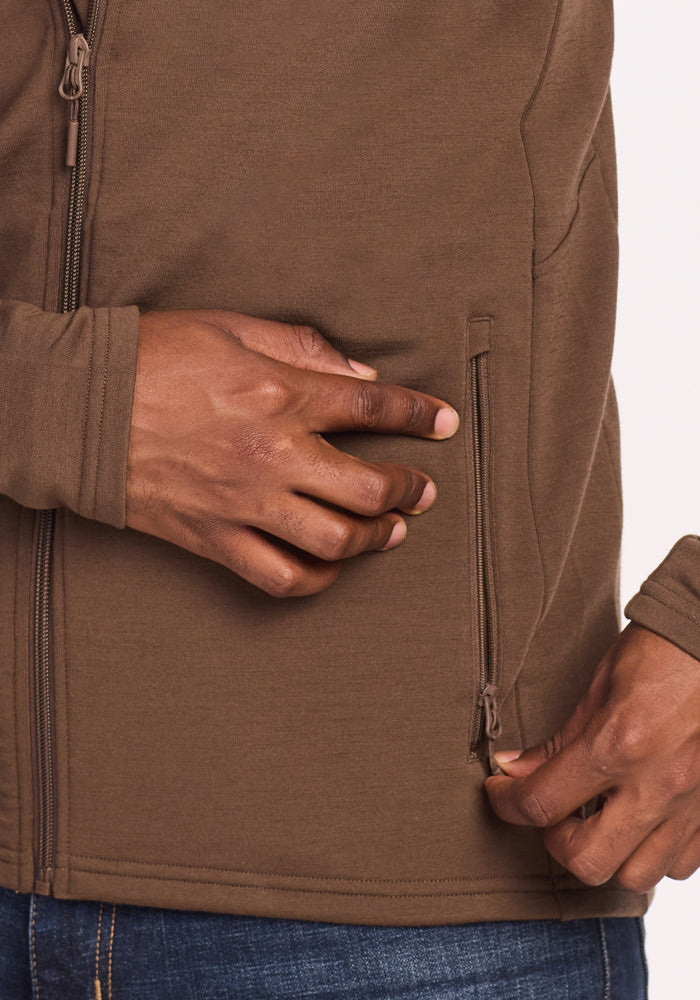 A close-up of a person wearing the Woolx Boulder Hoodie - Bourbon Oak and blue jeans, unzipping a side pocket on the hoodie with their right hand.