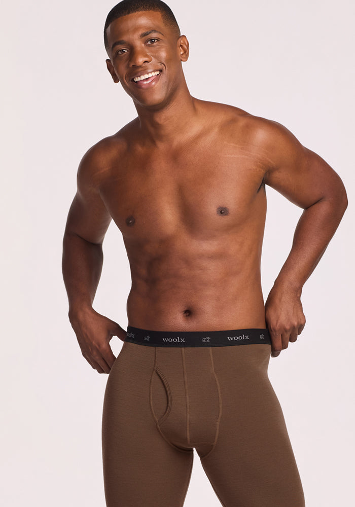 A smiling man stands against a light background, wearing only Woolx Backcountry Leggings - Bourbon Oak, featuring a black waistband with "woolx" in white letters.