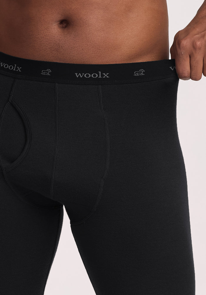 A person wears black Woolx Arctic Leggings, shown waist to mid-thigh. One hand pulls the waistband outward, revealing the "woolx" logo on these ultra-heavyweight thermal Merino Wool base layer leggings.