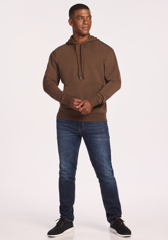 A man with short hair stands slightly turned, hands loosely clasped, wearing the Woolx Chase Hoodie in Bourbon Oak over blue jeans and black sneakers. The background is plain white.
