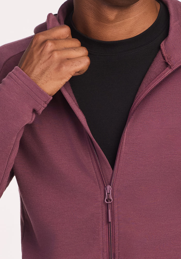 A person is shown from the shoulders down, unzipping the Woolx Boulder Hoodie - Grape Harvest with their right hand over a black shirt.