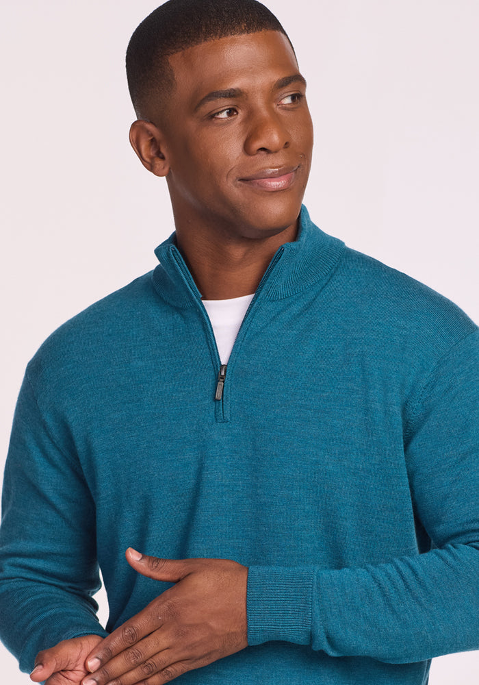 A man in a Woolx Enzo Quarter Zip - Harbor Teal, a midweight Merino wool layer, over a white shirt looks left and smiles slightly, one hand on his abdomen. The background is plain and light-colored.