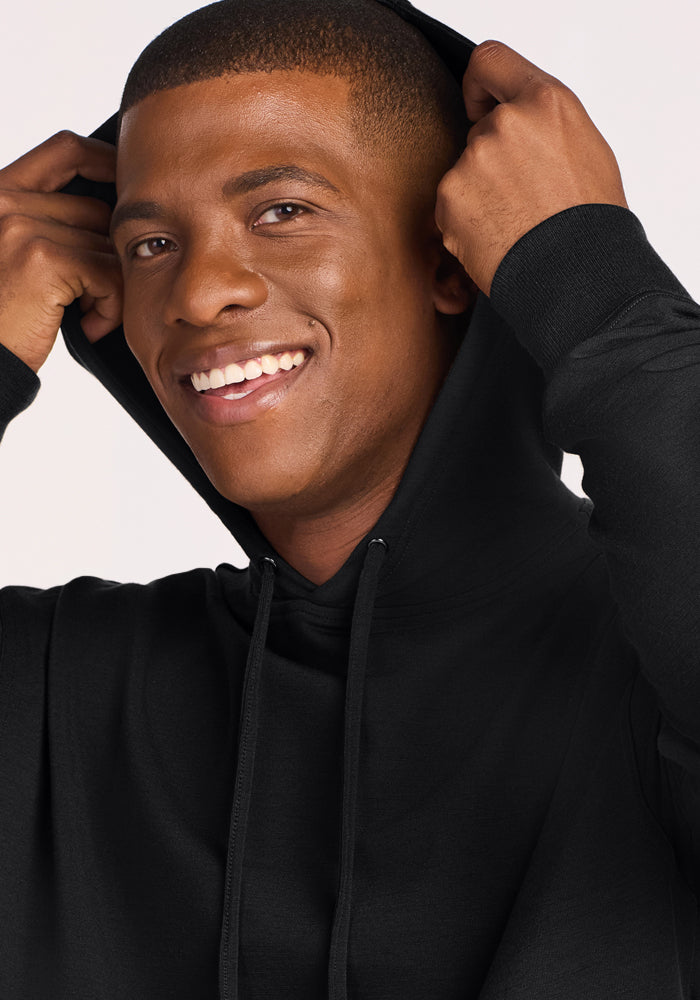 A smiling man with short hair pulls the hood over his head while wearing the Woolx Chase Hoodie in Black. He looks slightly to the side against a plain, light-colored background.