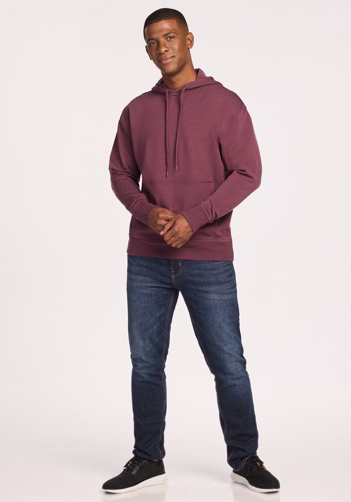 A man stands facing forward, wearing the Woolx Chase Hoodie in Grape Harvest, blue jeans, and black sneakers. He looks at the camera with a slight smile, hands loosely clasped, against a plain light background.