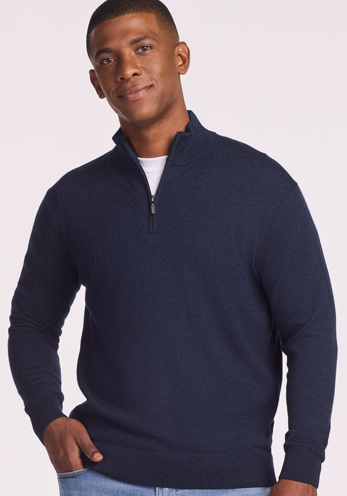 A man faces the camera, wearing the Woolx Enzo Quarter Zip - Storm Cloud over a white shirt and light blue jeans, one hand in his pocket and smiling slightly. This midweight layer brings comfort against a plain, light background.