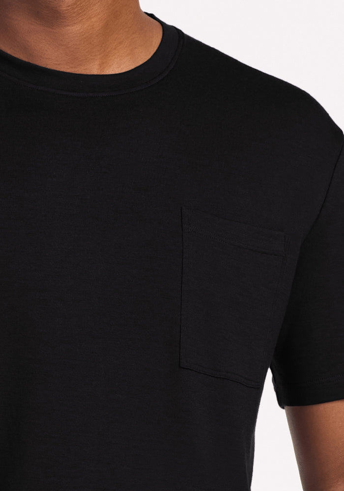 A close-up of a person wearing the Woolx Finn Top - Black, an odor-resistant Merino wool sleep tee with a left chest pocket, set against a plain light background. The person's head is partially out of frame.