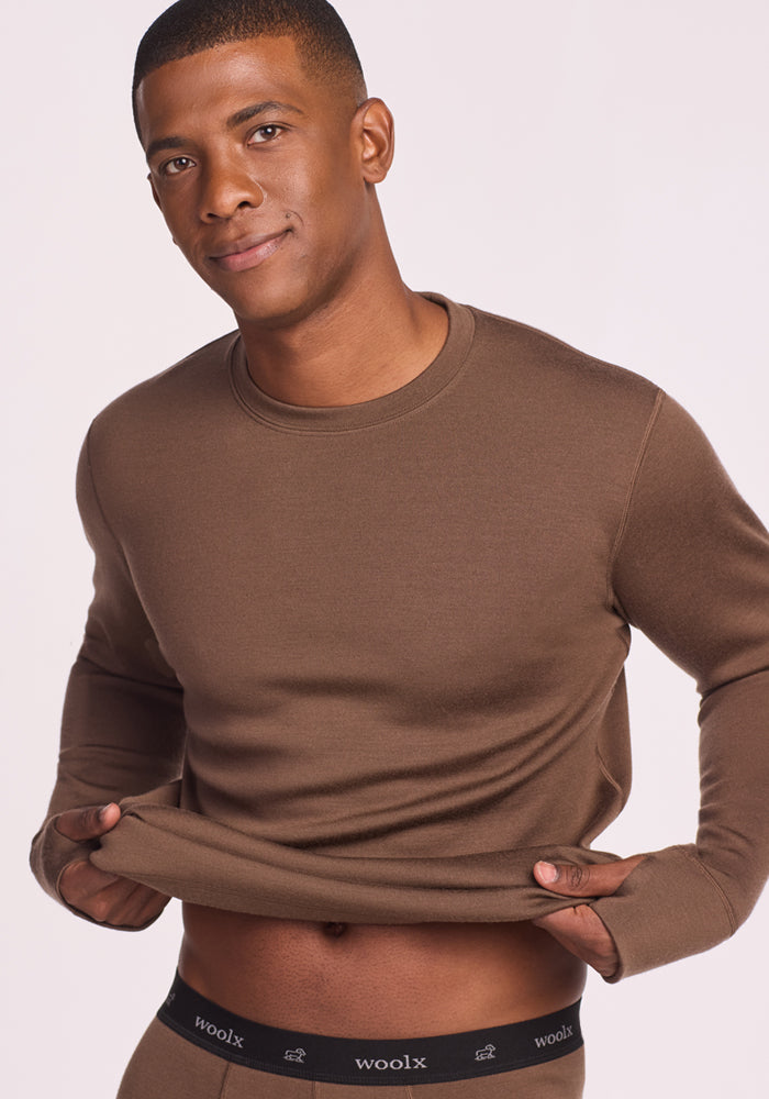 A man in a Woolx Glacier Long Sleeve Crew in Bourbon Oak lifts the hem to reveal black merino wool baselayer underwear with a "woolx" logo on the waistband. He smiles softly against a light background.