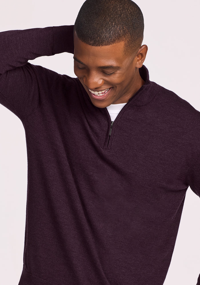 A smiling man wears the Woolx Enzo Quarter Zip in Deep Plum, standing with one hand behind his head and looking down. The plain, light background highlights his comfortable, temperature-regulating midweight sweater.