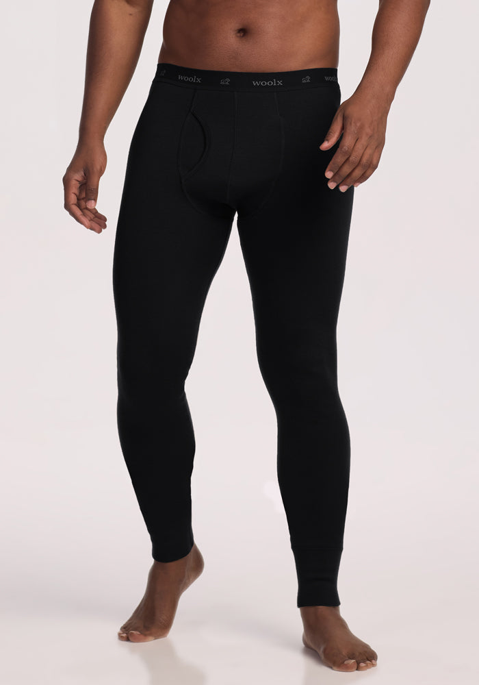 A man stands barefoot in Woolx Arctic Leggings - Black, ultra-heavyweight thermal long underwear featuring an elastic waistband with "woolx" in white, photographed against a plain light background.