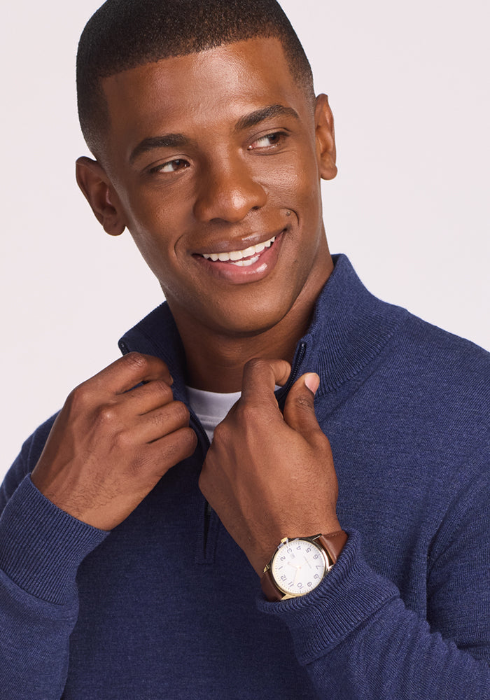 A smiling man wearing the Woolx Enzo Quarter Zip in Twilight Storm and a wristwatch adjusts the zipper of his midweight layer while looking to the side against a light background.