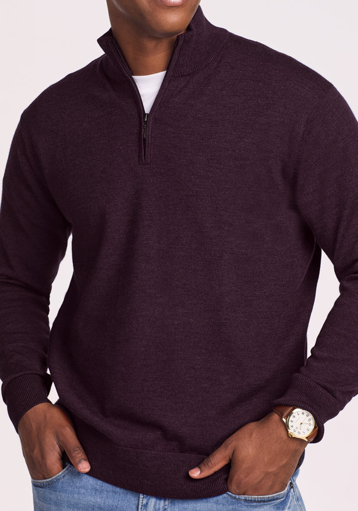 A man wears the Woolx Enzo Quarter Zip in Deep Plum over a white shirt, light blue jeans, and a gold watch, standing with hands in pockets. Its temperature-regulating fabric keeps him comfortable all day.