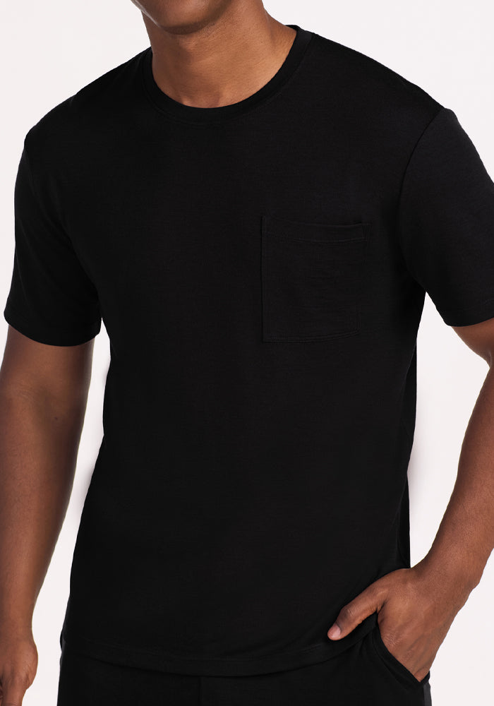 A person wearing the Woolx Finn Top - Black, an odor-resistant short-sleeve t-shirt with a chest pocket, stands against a white background. Only their upper body is visible, with one hand in their pocket.