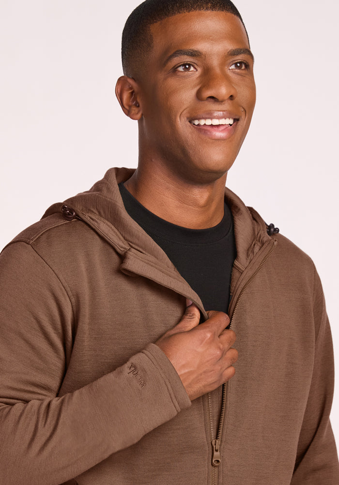 A smiling man wears the Woolx Grizzly Sweatshirt in Bourbon Oak over a black shirt, touching the zipper, and standing against a light background.