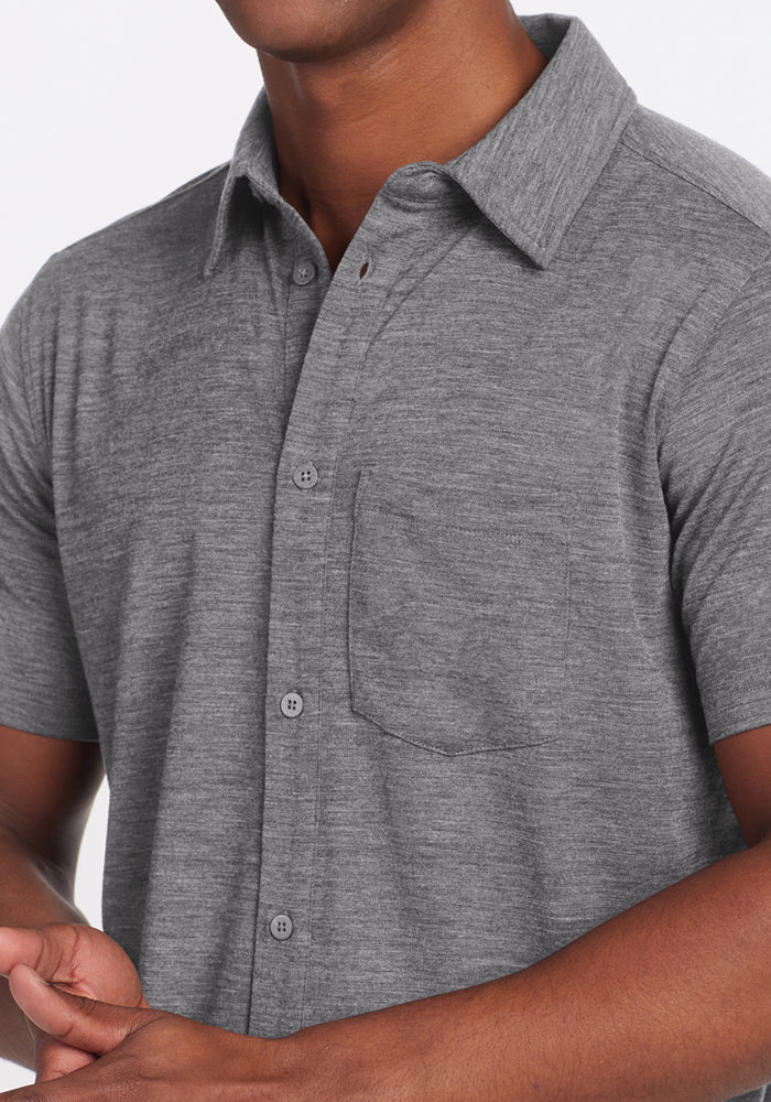A person wears the Woolx Endi Full Button Down Shirt in Graphite Heather, a lightweight short-sleeve style with a chest pocket. The image is cropped to show only their torso and arms, with the face not visible.