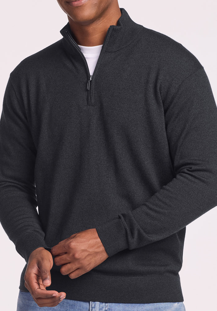 A person wears the Woolx Enzo Quarter Zip - Coal Stone, a midweight sweater, over a white shirt with light blue jeans. They stand clasping their hands against a plain, light-colored background—perfect for layering.