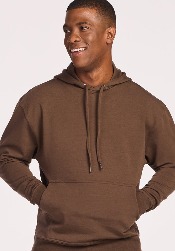 A smiling man wears the Woolx Chase Hoodie in Bourbon Oak, hands in the front pocket, standing against a plain light background.