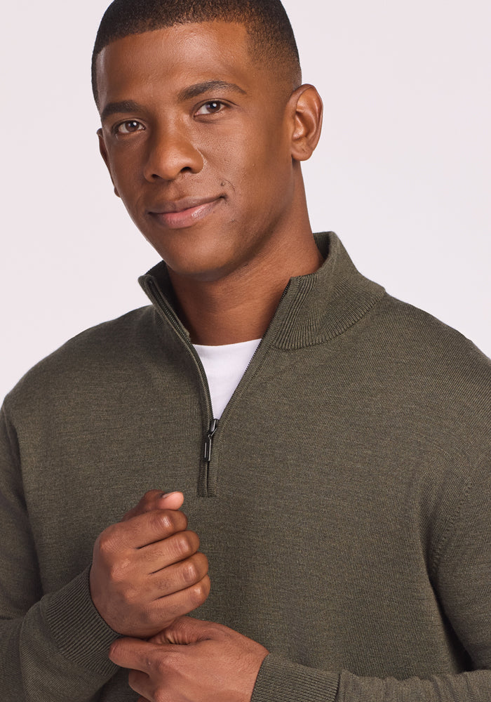 A man wearing the Woolx Enzo Quarter Zip in Pine Needle over a white shirt stands against a light background, smiling slightly at the camera with his hands clasped. This midweight layer is perfect for versatile comfort.