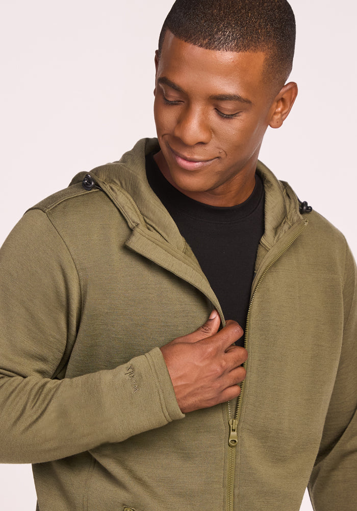 A man in the Woolx Grizzly Sweatshirt - Winter Moss, layered over a black shirt, holds the zipper and smiles slightly while looking down against a light background.