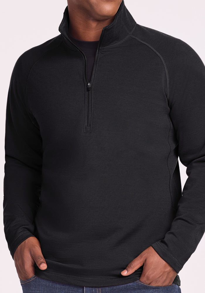 A person wearing the Woolx Blizzard 1/4 Zip in black stands against a light background, hands in blue jean pockets, with their face not visible.