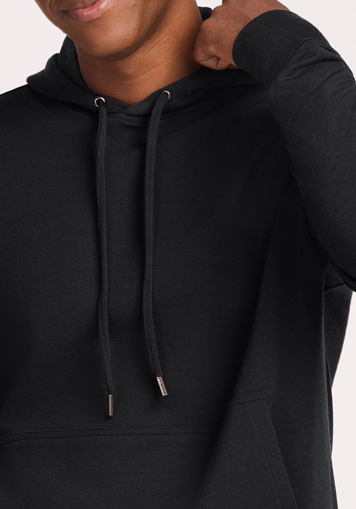A person wearing the Woolx Chase Hoodie - Black, shown from shoulders to mouth, is slightly smiling while holding the hood with one hand.