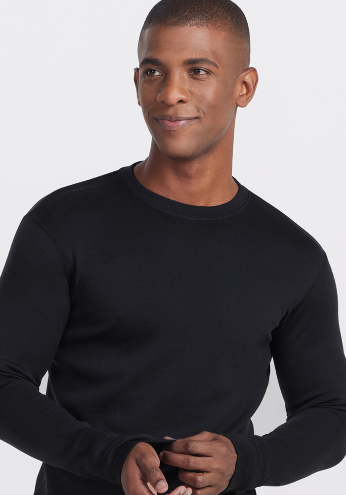 A man wearing the Woolx Glacier Long Sleeve Crew in black stands against a plain light background, looking slightly to his left and smiling softly.