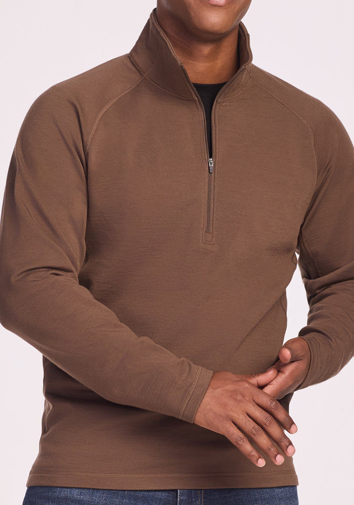 A person wears the Woolx Blizzard 1/4 Zip in Bourbon Oak with blue jeans, standing with clasped hands and facing forward. Their head is partially visible against a plain, light-colored background.