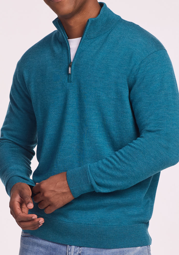 A man wears the Woolx Enzo Quarter Zip in Harbor Teal over a white shirt and light blue jeans, standing against a plain background with hands clasped—a midweight layer for effortless style.