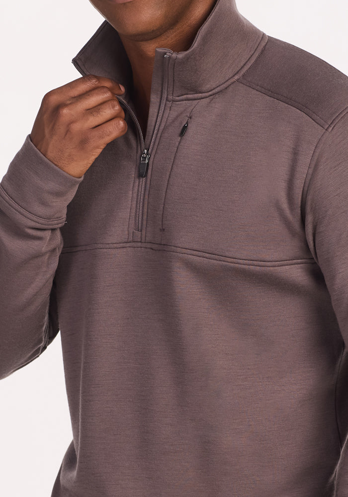 A person wears the Woolx Mason 1/4 Zip in Plum Truffle, pulling up the zippered high collar with one hand. Their face is partially out of frame.
