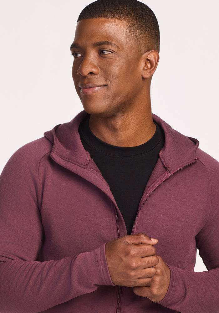 A man wearing the Woolx Boulder Hoodie in Grape Harvest over a black shirt stands against a plain background, looking to the side with a slight smile and clasping his hands together.