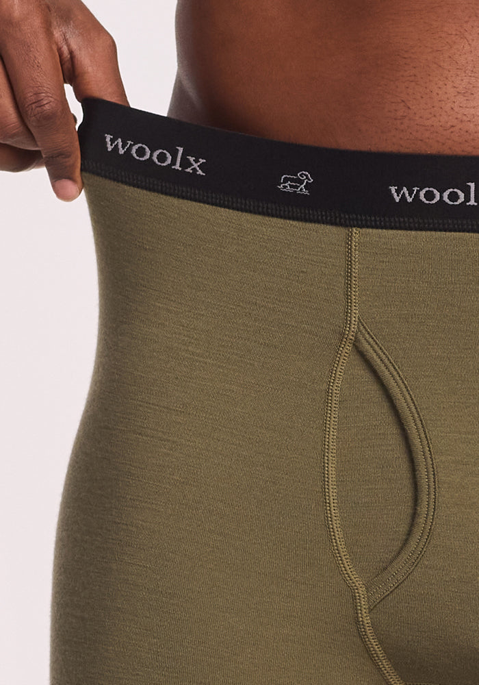A person pulls at the waistband of Woolx Backcountry Leggings in Winter Moss, revealing the black elastic band with the Woolx logo. The image focuses on the waist and hip area.