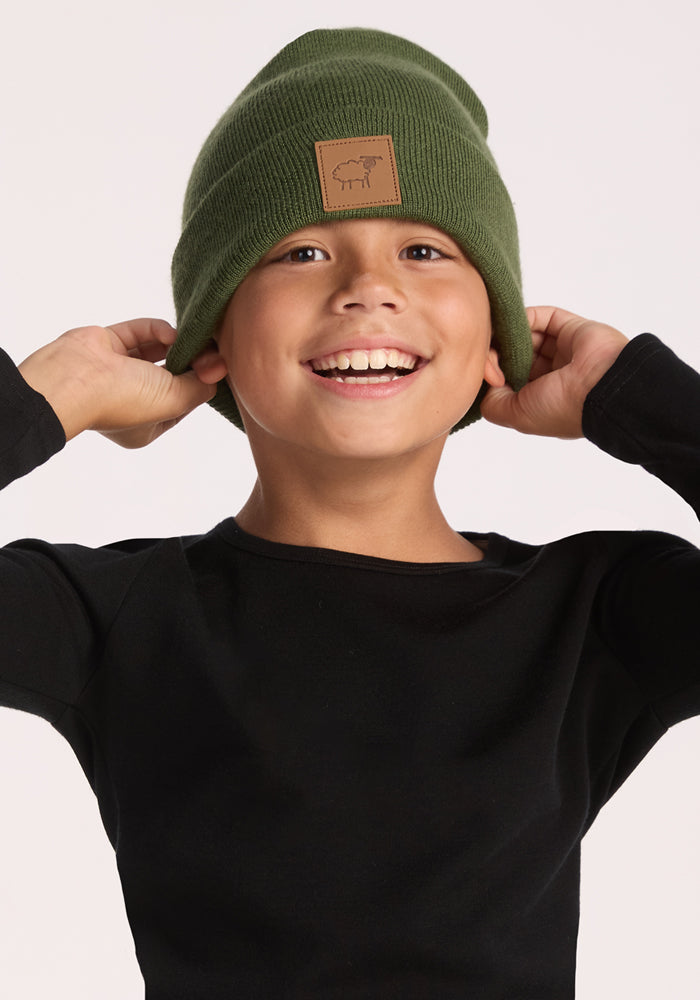 A smiling child wearing a Woolx Kids Baylor Beanie - Turtle Shell and a black long-sleeve shirt adjusts the beanie with both hands against a plain light background.
