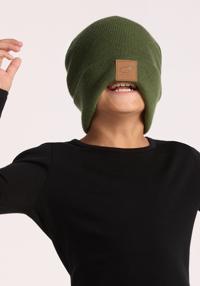 A child in a green Woolx Kids Baylor Beanie - Turtle Shell smiles with teeth showing, arms raised. The ultra-soft beanie and black shirt create a playful look against the light background.
