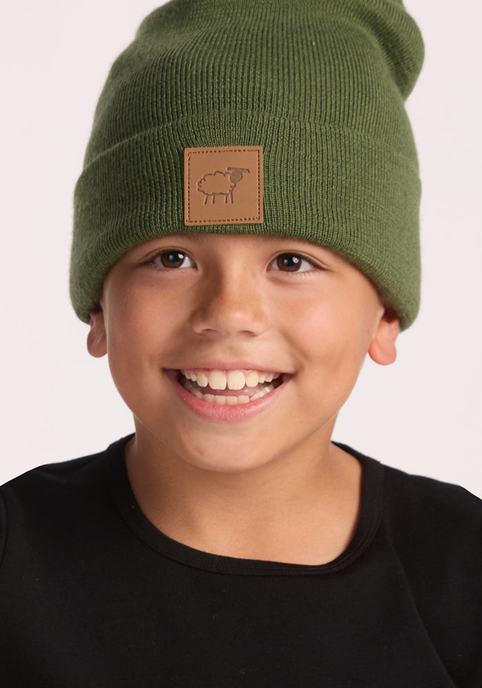 A smiling child wears a Woolx Kids Baylor Beanie - Turtle Shell, featuring a brown patch with a simple sheep illustration, and a black shirt, against a plain light background.