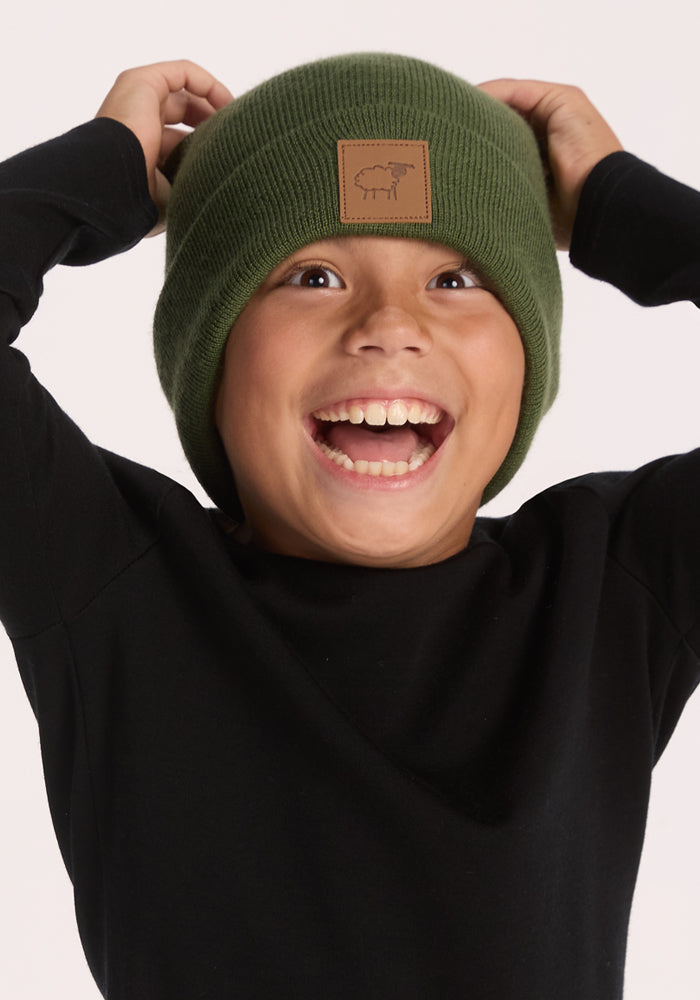 A smiling child wearing a Woolx Kids Baylor Beanie - Turtle Shell with a sheep logo and a black long-sleeve shirt, adjusting the ultra-soft, stretch-fit Merino wool beanie.