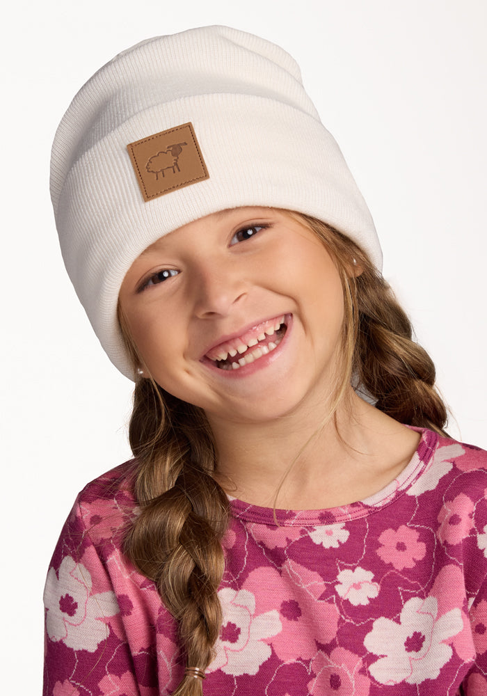 A young girl with long brown hair in a braid smiles brightly, wearing the Woolx Kids Baylor Beanie in Whipped Cream and a pink floral shirt. The beanie features a brown sheep patch. The background is plain white.