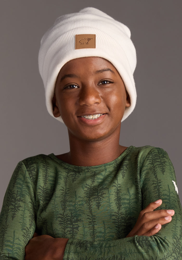A smiling child with medium skin tone wears the Woolx Kids Baylor Beanie - Whipped Cream, featuring a brown patch and full ear coverage, plus a green, tree-patterned long-sleeve shirt, standing arms crossed against a plain gray background.