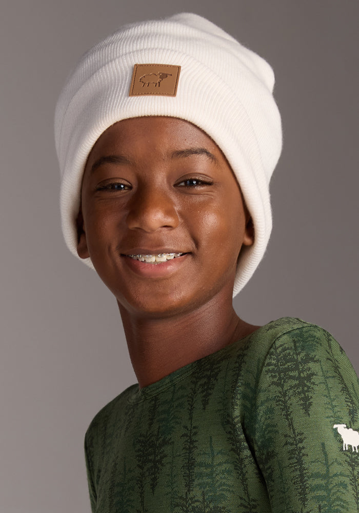 A smiling child wears the Woolx Kids Baylor Beanie in Whipped Cream, paired with a green shirt adorned with a tree pattern and a small white animal on the sleeve, posing against a neutral gray background.
