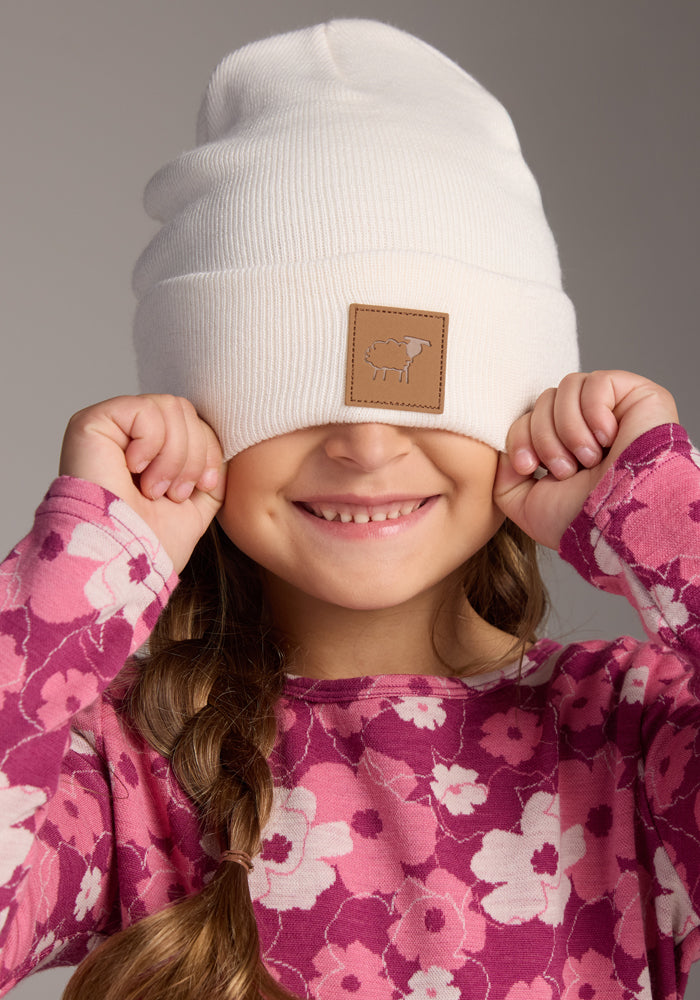 A smiling child with braided hair pulls the Woolx Kids Baylor Beanie in Whipped Cream, made from soft Merino wool, over her eyes. The beanie offers full ear coverage and has a brown patch featuring a small sheep design on the front.