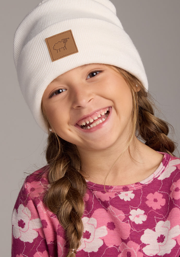 A young girl with long braids wears the Woolx Kids Baylor Beanie in Whipped Cream, featuring a sheep patch for full ear coverage, and a pink floral shirt as she smiles brightly against a plain background.
