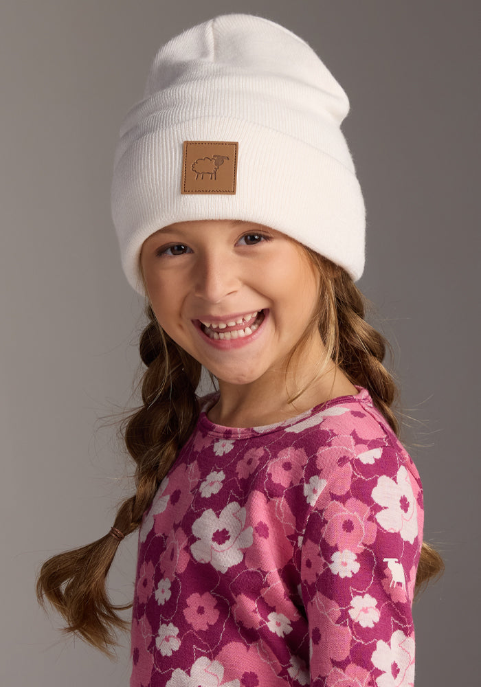A smiling young girl with two braids wears the Woolx Kids Baylor Beanie in Whipped Cream, featuring full ear coverage and a brown patch, paired with a pink floral shirt, posing against a plain gray background.